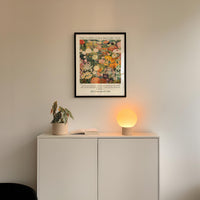 a white cabinet with a potted plant and a lamp on top, set against a white wall. Above the cabinet, there is a framed poster with a colorful floral design.