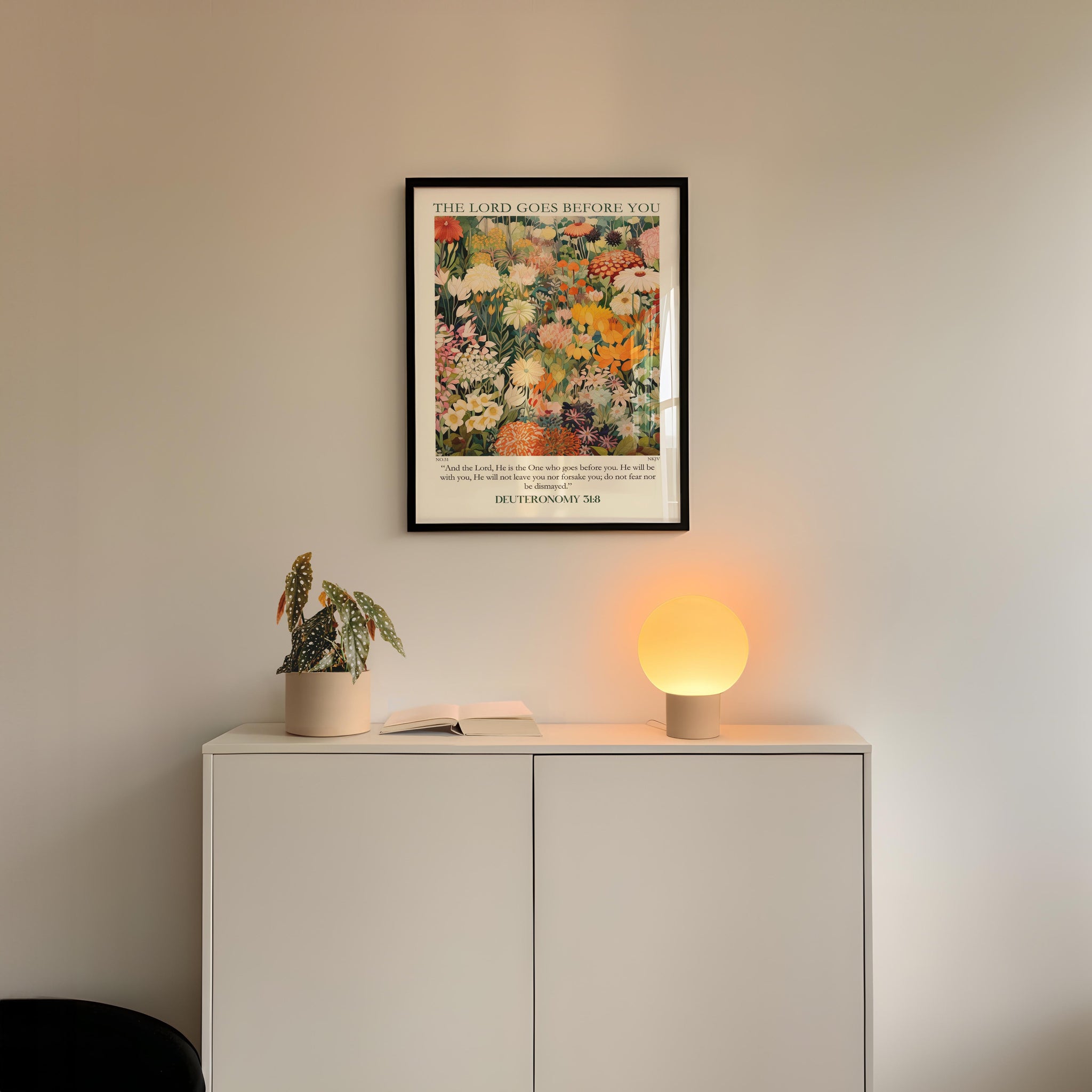 a white cabinet with a potted plant and a lamp on top, set against a white wall. Above the cabinet, there is a framed poster with a colorful floral design.