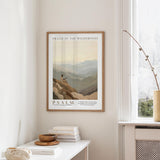 a framed poster of a mountain landscape hanging on a wall, with a window and a small table with books and a bowl on it below the poster.