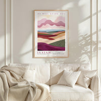 a framed poster of a landscape with mountains and hills, hanging on a wall above a white couch with a blanket draped over it.
