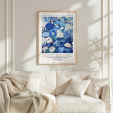 a framed poster of a blue and white floral design hanging on a white wall above a white couch with a blanket draped over it.