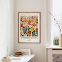 a framed poster of a vibrant floral design hanging on a wall, with a window and a small table with books and a bowl in the foreground.