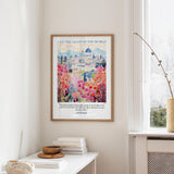 a framed poster of a colorful, artistic depiction of a cityscape, with a quote from the Bible above it. The poster is hanging on a wall next to a window, and there is a small table with books and a vase on it.