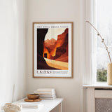 a framed poster of a desert landscape with mountains, hanging on a wall in a room with a white table and a window.