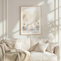 a framed poster of a serene landscape with clouds and a golden crown, hanging on a white wall above a white couch with a blanket draped over it.