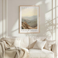 a framed poster of a person sitting on a mountain, with a serene landscape in the background. The poster is titled "PRAISE IN THE WILDERNESS" and includes a Bible verse from the book of Psalms. The poster is hung on a white wall above a white couch, which has a blanket draped over it.
