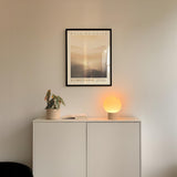 a minimalist interior design with a white cabinet, a potted plant, and a lamp on a shelf. Above the cabinet, there is a framed poster with the text "WALK BY FAITH" and "2 ZORINTHANS" written on it.