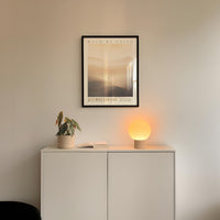 a minimalist interior design with a white cabinet, a potted plant, and a lamp on a shelf. Above the cabinet, there is a framed poster with the text "WALK BY FAITH" and "2 ZORINTHANS" written on it.