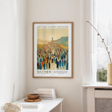 a framed poster of a large crowd of people, with the text "JESUS fed the five thousand" and "MATTHEW 1419 New Testament" below it. The poster is hanging on a wall, and there is a small table with a stack of books and a vase on it.