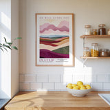 A framed poster of a landscape with mountains and hills hangs on a wall above a wooden countertop with a bowl of lemons.