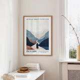 a framed poster of a mountain landscape with a quote from Proverbs, hanging on a wall in a room with a white table and a vase of flowers.