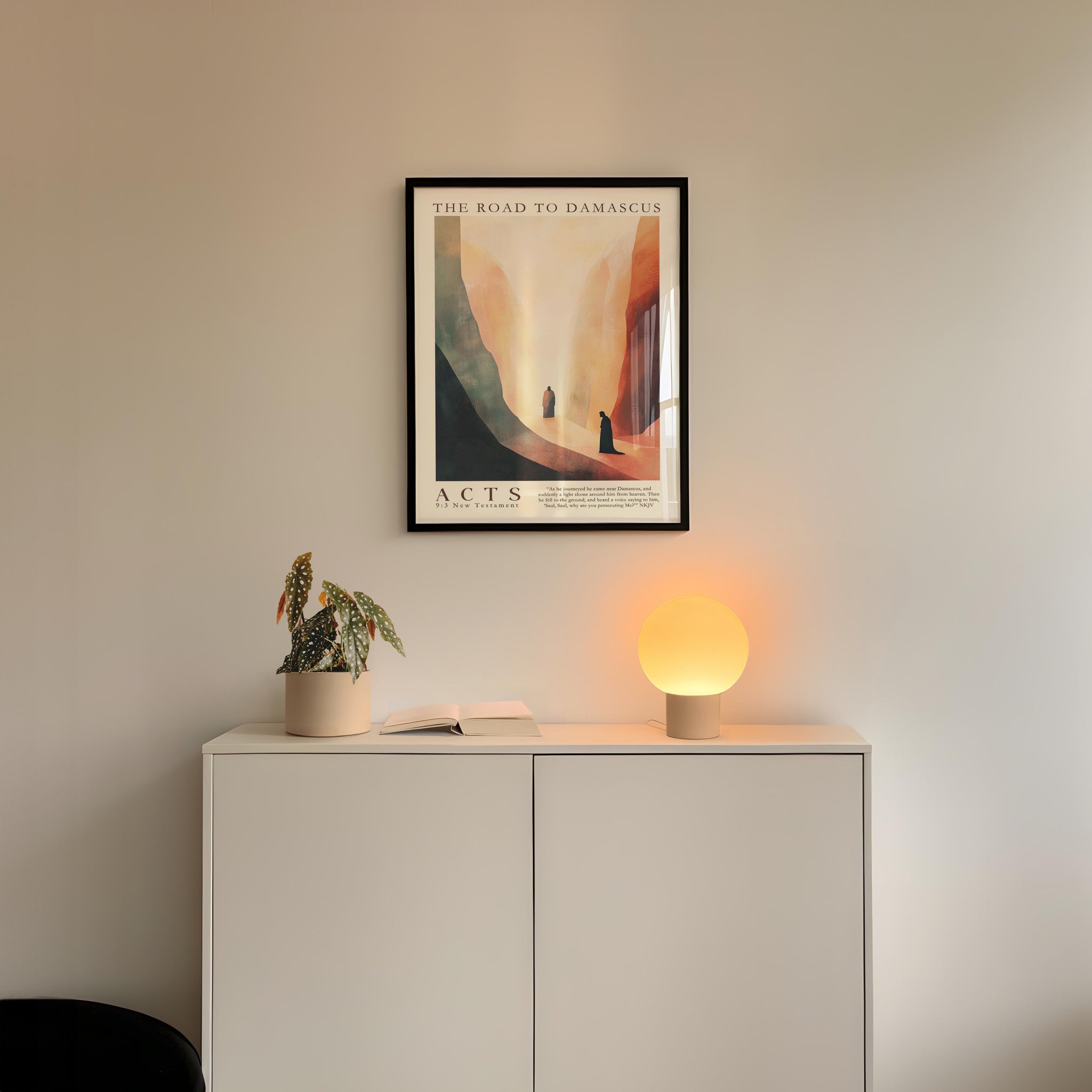 a minimalist interior design with a framed poster of a desert landscape hanging on a white wall above a white cabinet. The cabinet has a plant and a lamp on top of it, creating a cozy and inviting atmosphere.