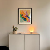 a white cabinet with a plant and a lamp on top, and a framed poster hanging on the wall above the cabinet.