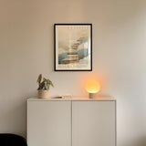 a white cabinet with a plant and a lamp on top, and a framed poster of a staircase above it.