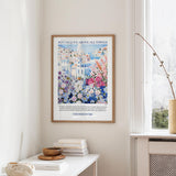 a framed poster of a colorful floral scene hanging on a wall, with a white table and a vase of flowers in the foreground.