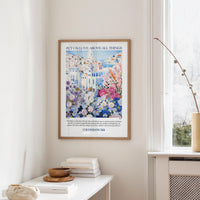 a framed poster of a colorful floral scene hanging on a wall, with a white table and a vase of flowers in the foreground.