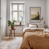 A cozy bedroom with a large bed, a wooden nightstand, and a potted plant. The room has a neutral color scheme with white walls and a wooden floor. A framed poster on the wall adds a decorative touch, and a window with a plant on the sill brings a touch of nature into the space.