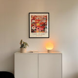 a white cabinet with a potted plant and a lamp on top, set against a white wall. Above the cabinet, there is a framed poster with a floral design.