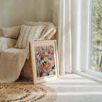 a framed poster of a colorful floral design, placed on a wooden floor next to a window with white curtains.