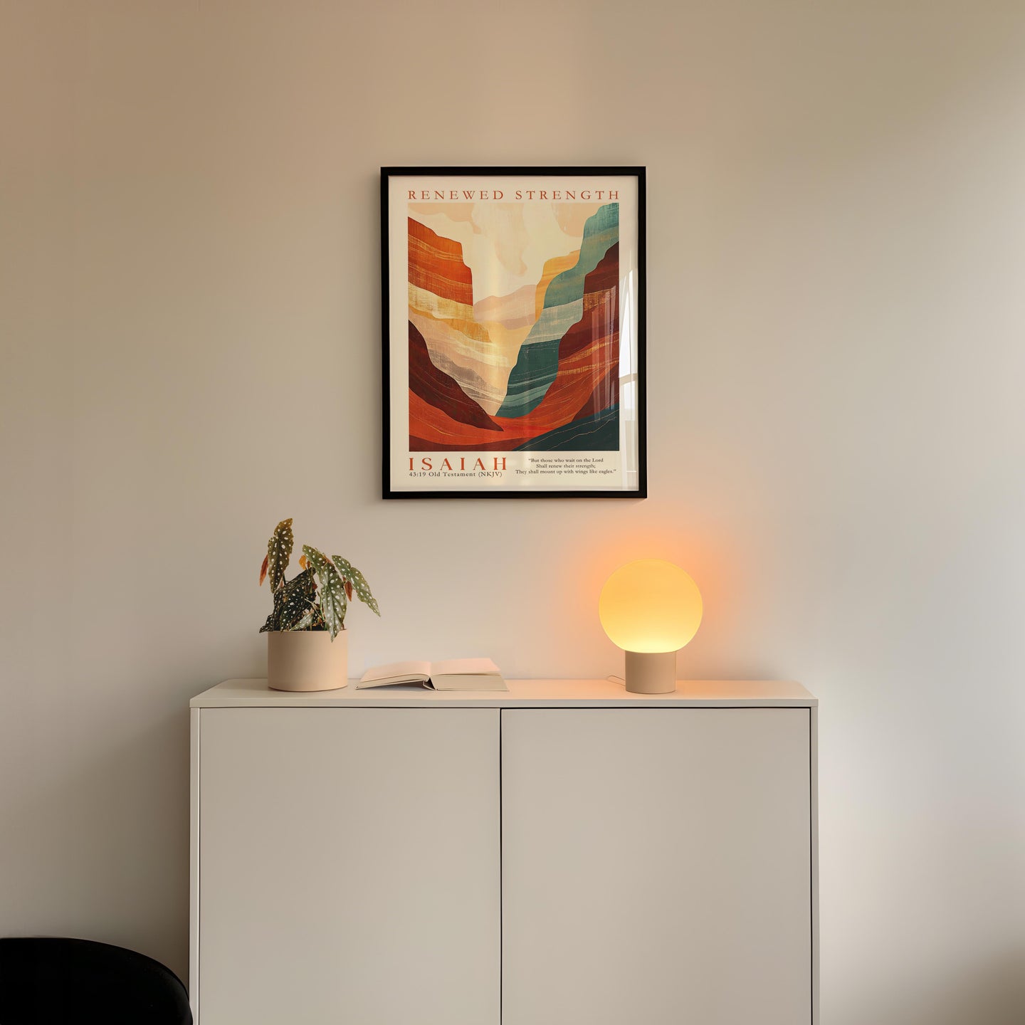 a white cabinet with a potted plant and a lamp on top, and a framed poster of a mountain landscape hanging on the wall above it.
