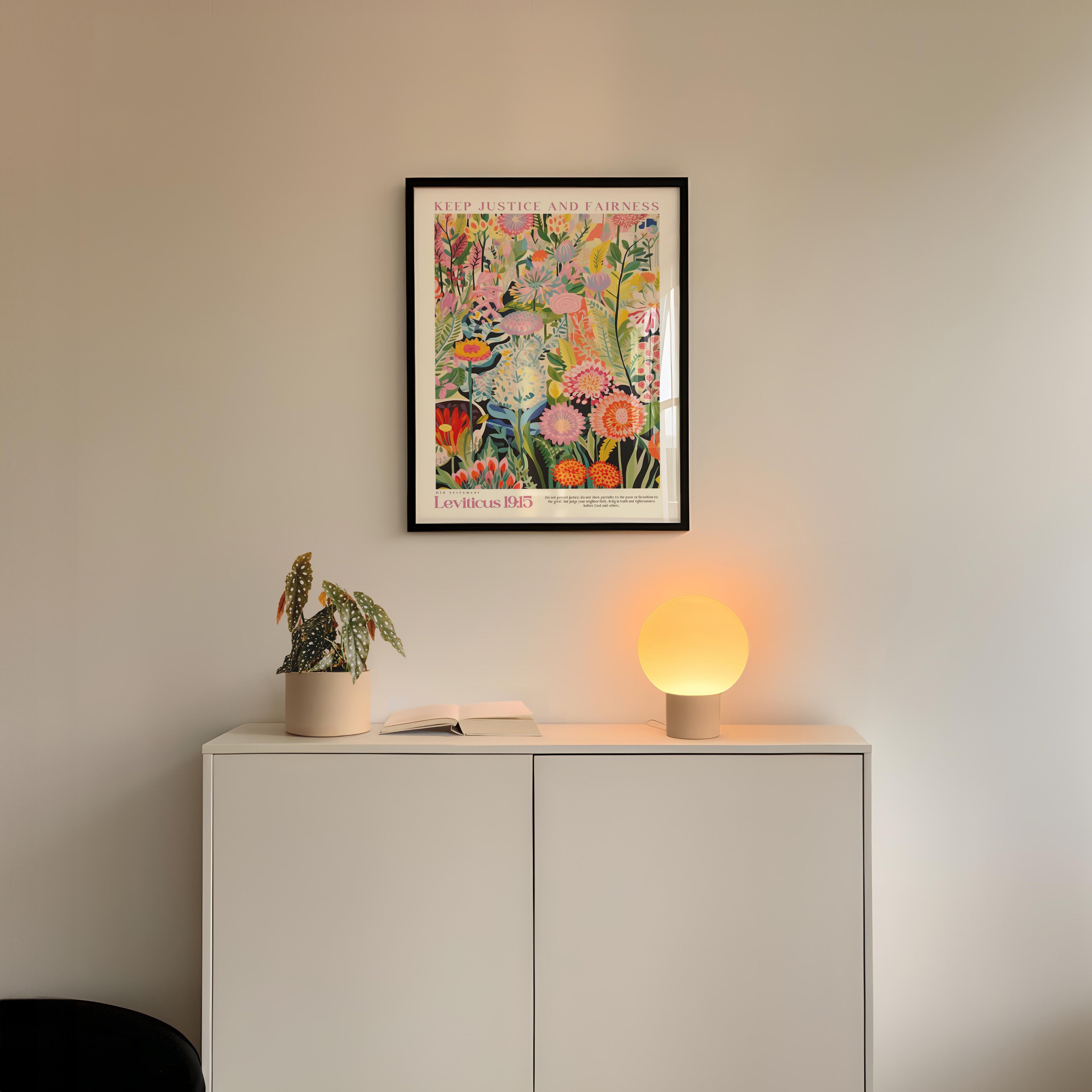a white cabinet with a potted plant and a lamp on top, set against a white wall. Above the cabinet, there is a framed poster with a colorful floral design.