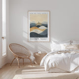 A bedroom with a large bed, a woven chair, and a framed poster of mountains on the wall.