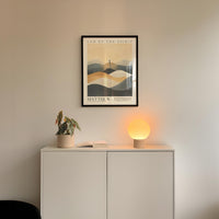 a minimalist interior design with a framed poster of a landscape, a potted plant, and a small lamp on a white cabinet.