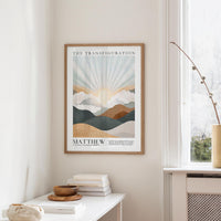 a framed poster of a mountain landscape with a sunburst design, hanging on a wall above a white table with a stack of books and a small vase.
