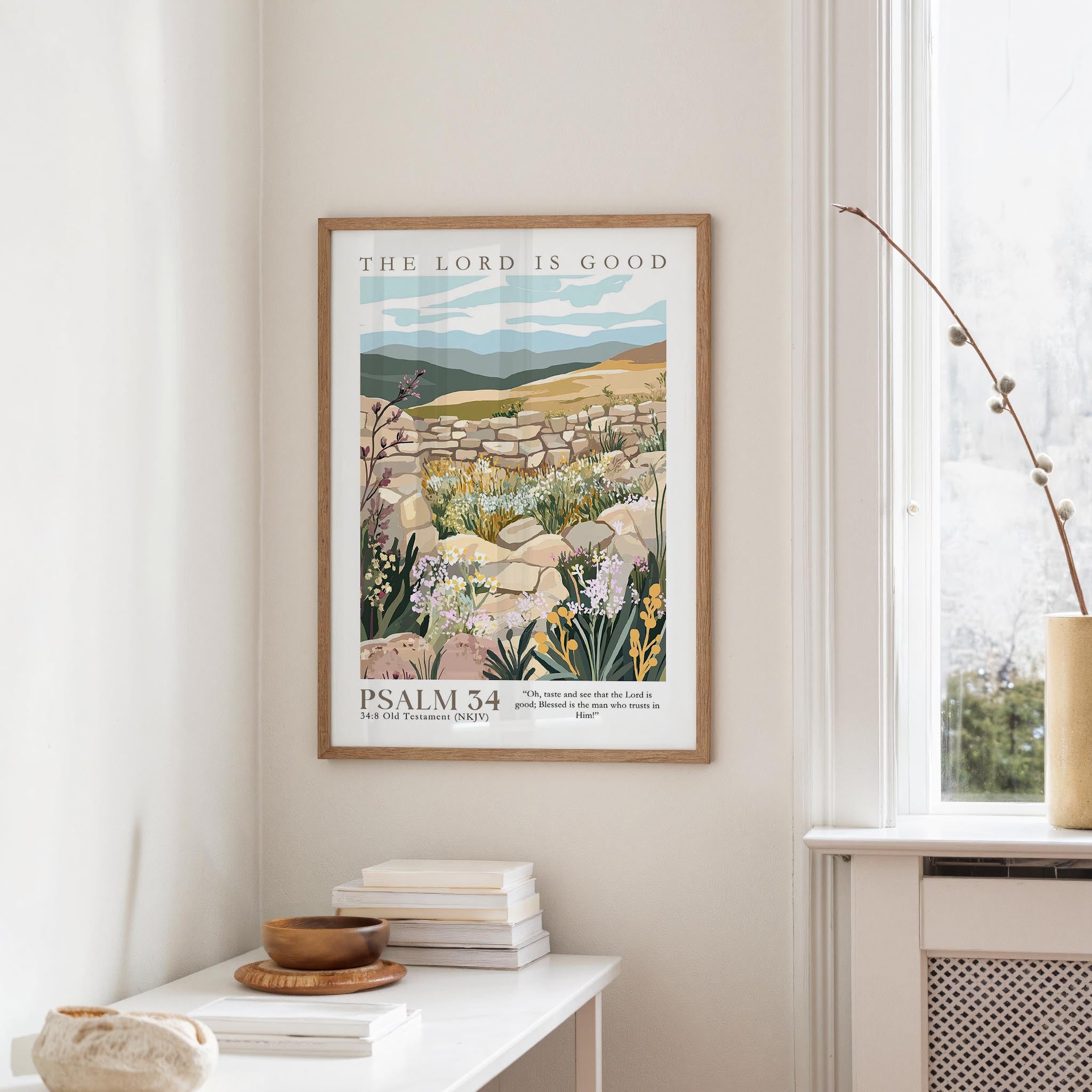 a framed poster of a pastoral landscape with a bird perched on a rock, hanging on a wall in a room with a window and a small table with books and a bowl.