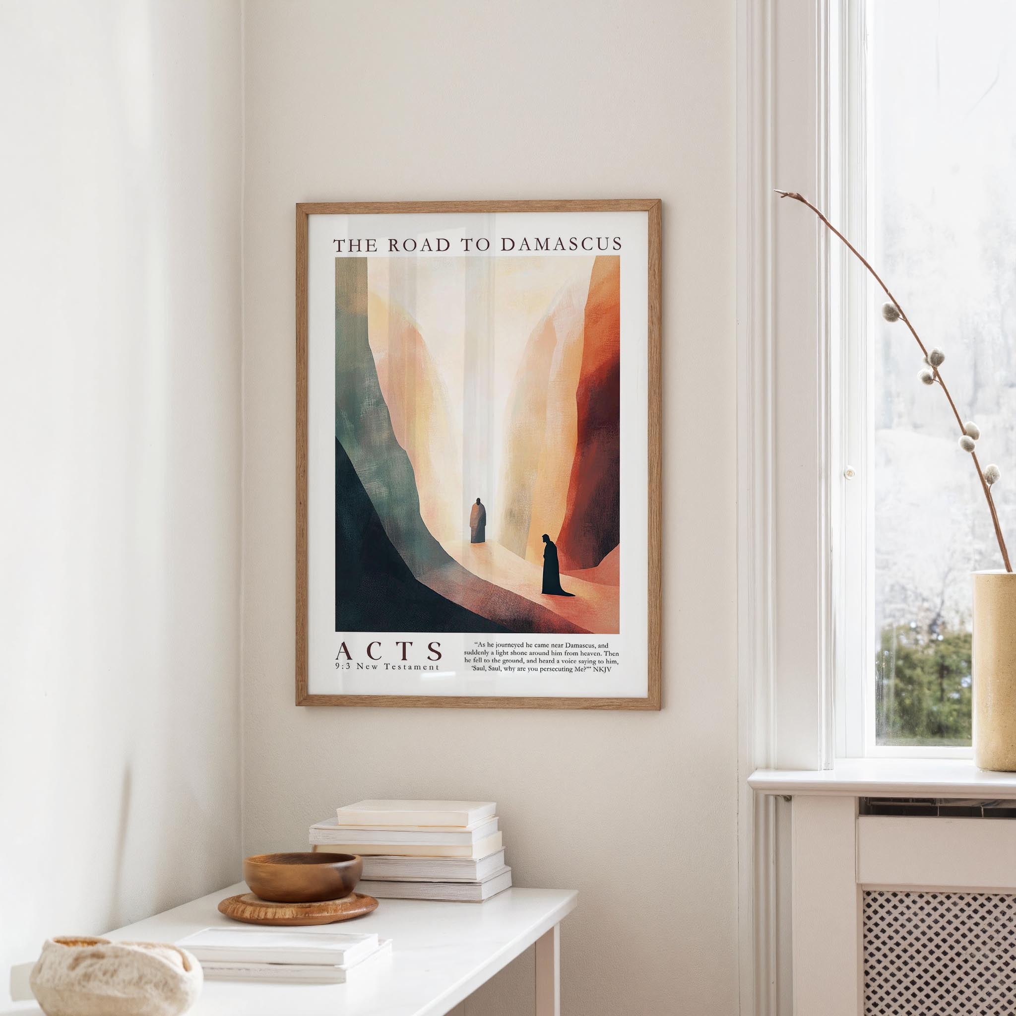 a framed poster of a desert landscape with two figures walking through it, hanging on a wall in a room with a white table and a window.