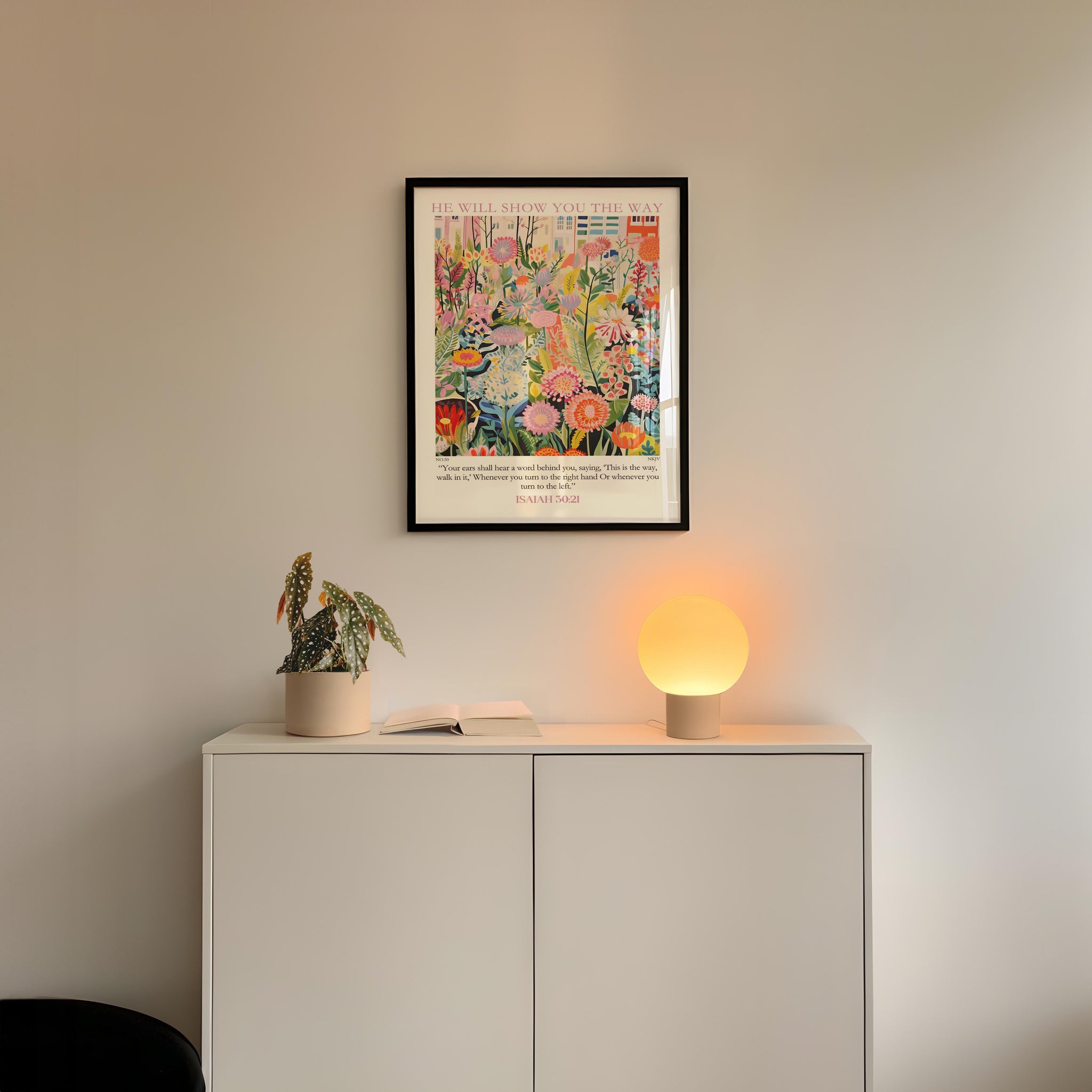 a white cabinet with a plant and a lamp on top, and a framed colorful painting hanging on the wall above it.