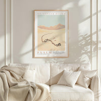 a framed poster of a desert landscape with mountains in the background, hanging on a wall above a white couch with a blanket draped over it.
