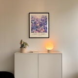 a white cabinet with a potted plant and a lamp on top, set against a white wall. Above the cabinet, there is a framed poster with a floral design.