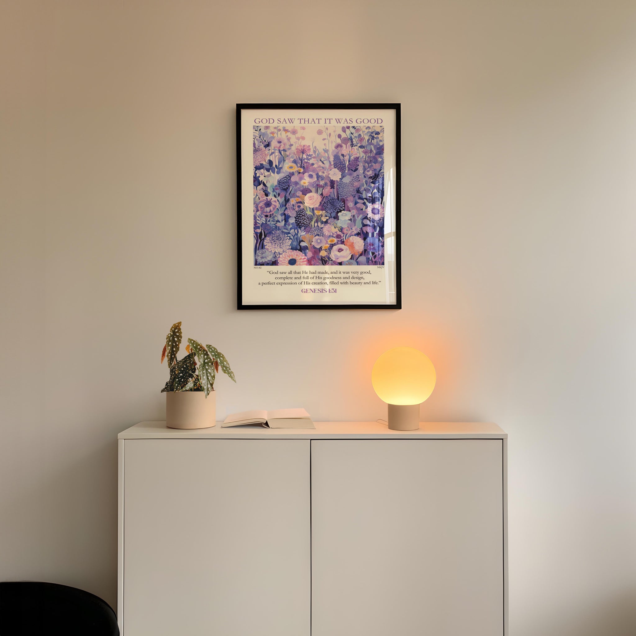 a white cabinet with a potted plant and a lamp on top, set against a white wall. Above the cabinet, there is a framed poster with a floral design.