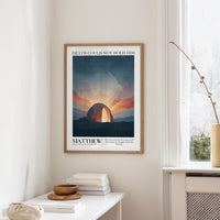 a framed poster of a sunset over a desert landscape, hanging on a wall in a room with a white table and a vase of flowers.
