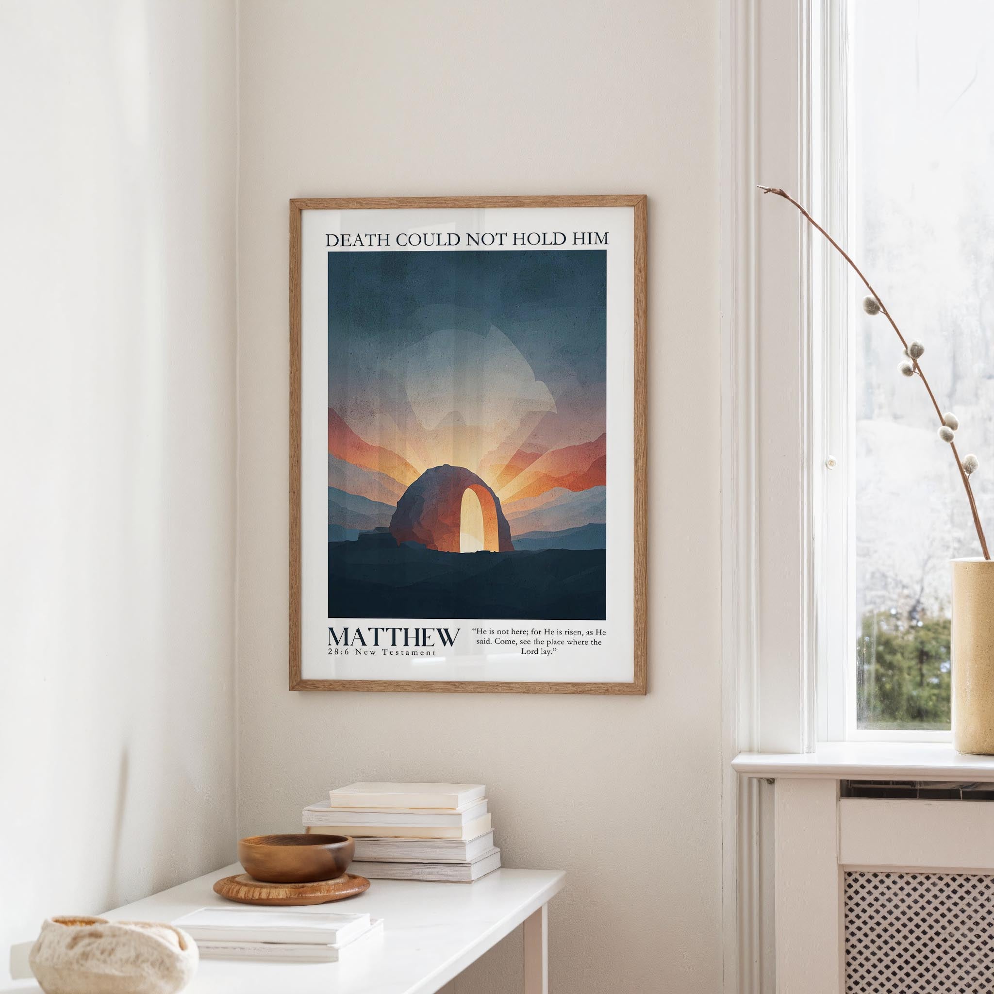 a framed poster of a sunset over a desert landscape, hanging on a wall in a room with a white table and a vase of flowers.