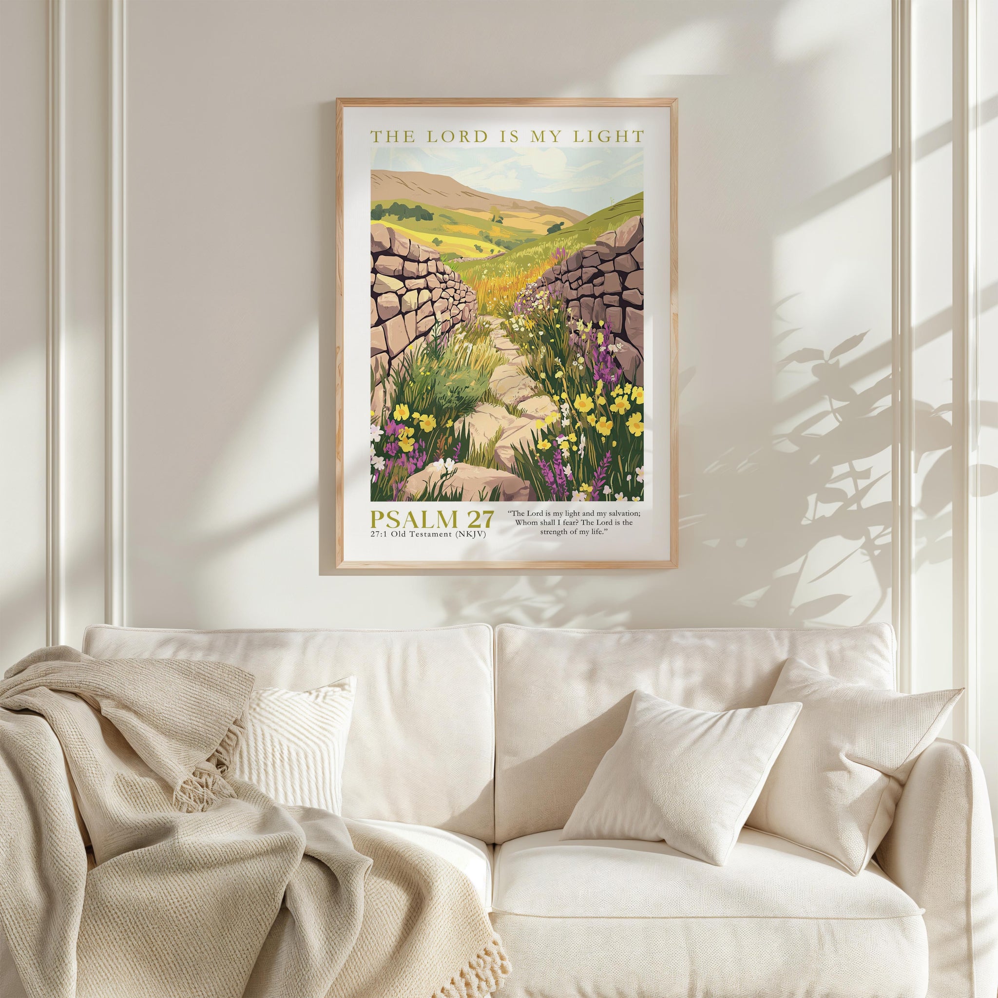 a framed poster of a stone path leading through a field of flowers, with the text "THE LORD IS MY LIGHT" and "PSALM 27" below it. The poster is hanging on a white wall above a white couch, which has a blanket draped over it.