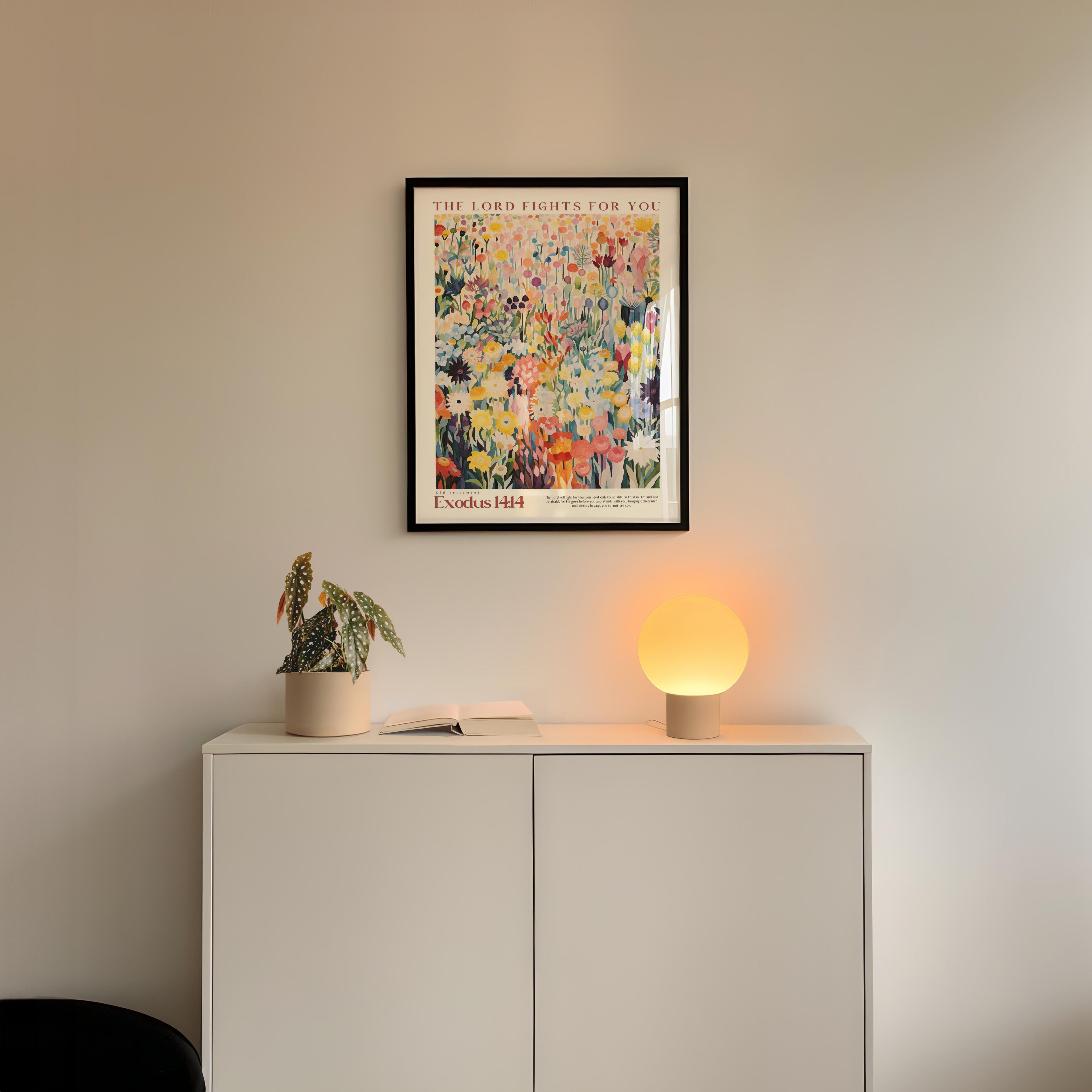 a white cabinet with a potted plant and a lamp on top, set against a white wall. Above the cabinet, there is a framed poster with a colorful abstract painting.