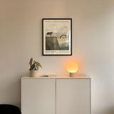 a minimalist interior design with a white cabinet, a potted plant, and a lamp on a shelf. Above the cabinet, there is a framed poster depicting a house on a cliff.