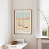 a framed poster of a desert landscape with mountains in the background, hanging on a wall above a white table with a stack of books and a small vase.