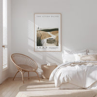 A bedroom with a bed, a woven chair, and a framed poster of a rural landscape with a well in the background.