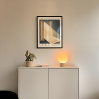 a minimalist interior design with a framed poster of a staircase and a small plant on a white cabinet. The poster has the text "THE BOOK OF LIFE" and "LUKJ" written on it. Below the poster, there is a small plant in a white pot, and a small lamp is placed on the cabinet.