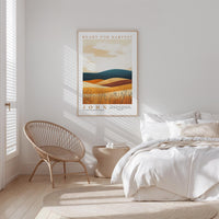 a serene bedroom scene with a bed, a woven chair, and a framed poster of a desert landscape.