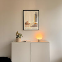a minimalist interior design with a framed poster of a building, a potted plant, and a small lamp on a white cabinet.
