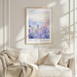 a framed poster of a serene landscape with a person standing in the distance, surrounded by purple flowers, hanging on a white wall above a beige couch with pillows.