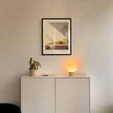 a minimalist interior design with a framed poster of a loaf of bread and a small plant on a white cabinet. Above the cabinet, there is a lamp with a glowing light, creating a warm and cozy atmosphere.