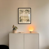 a white cabinet with a plant and a lamp on top, and a framed poster of a landscape hanging on the wall above it.