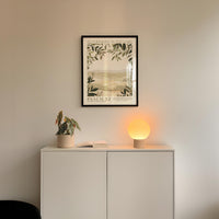 a white cabinet with a plant and a lamp on top, and a framed poster of a landscape hanging on the wall above it.
