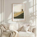 a framed poster of a desert landscape with a small tower on a white wall above a white couch with a blanket draped over it.