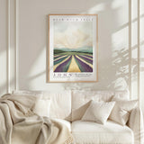 a framed poster of a landscape with a field of fruit trees, hanging on a wall above a white couch with a blanket draped over it.